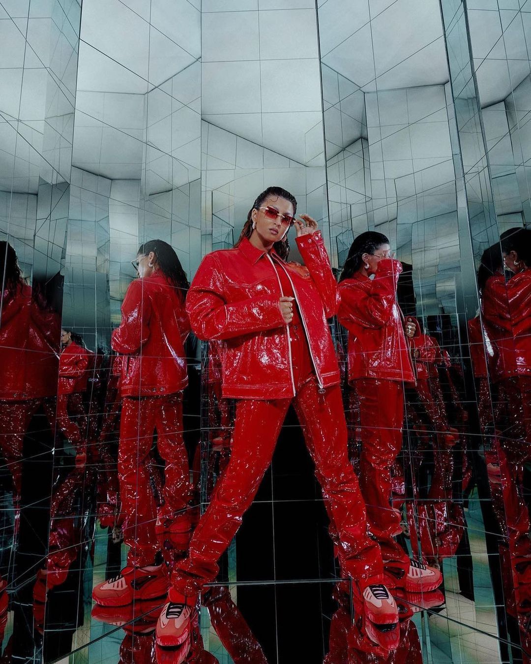A woman in a red patent leather outfit poses in a room of mirrors, creating multiple reflections. The image emphasizes fashion and reflective space.