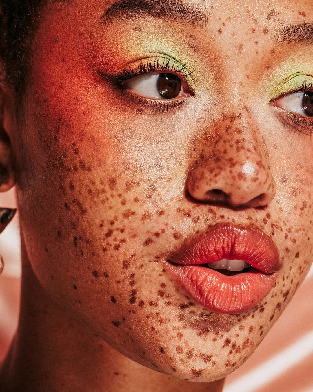 Close-up studio shot of a young woman with freckles and colorful makeup, looking off-camera, with a vibrant background, highlighting beauty and fashion.
