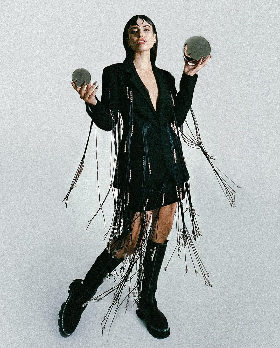 A woman in a black jacket dress with fringe poses in a photo studio, holding metallic, reflective round objects in her hands.