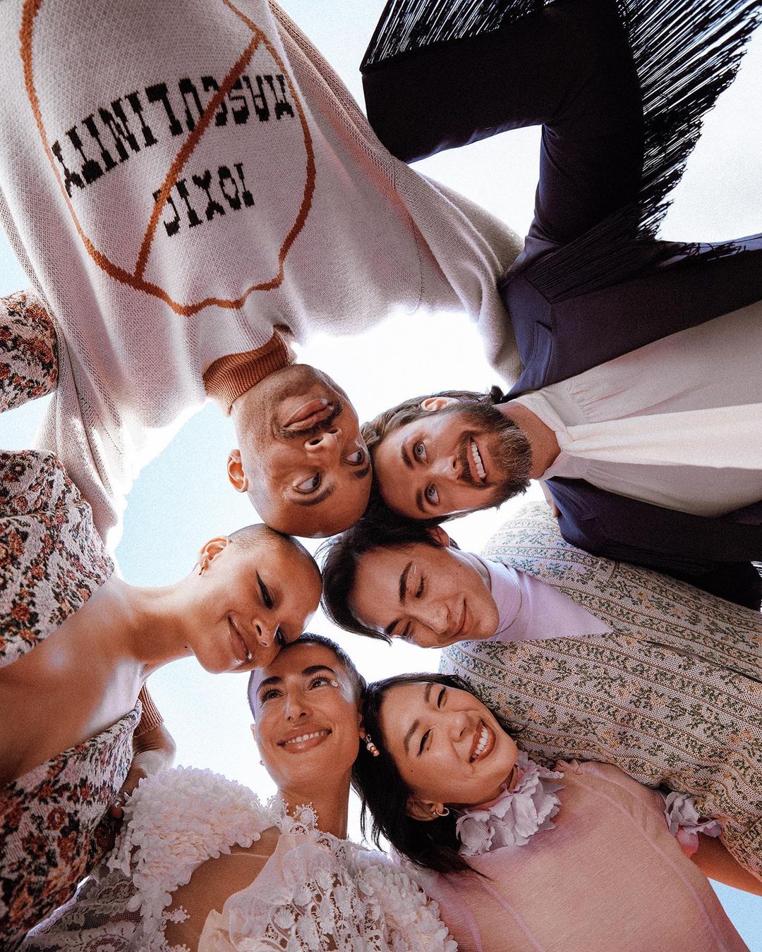 Group portrait of five smiling adults in a huddle, showcasing diverse fashion and unity against a light sky backdrop.