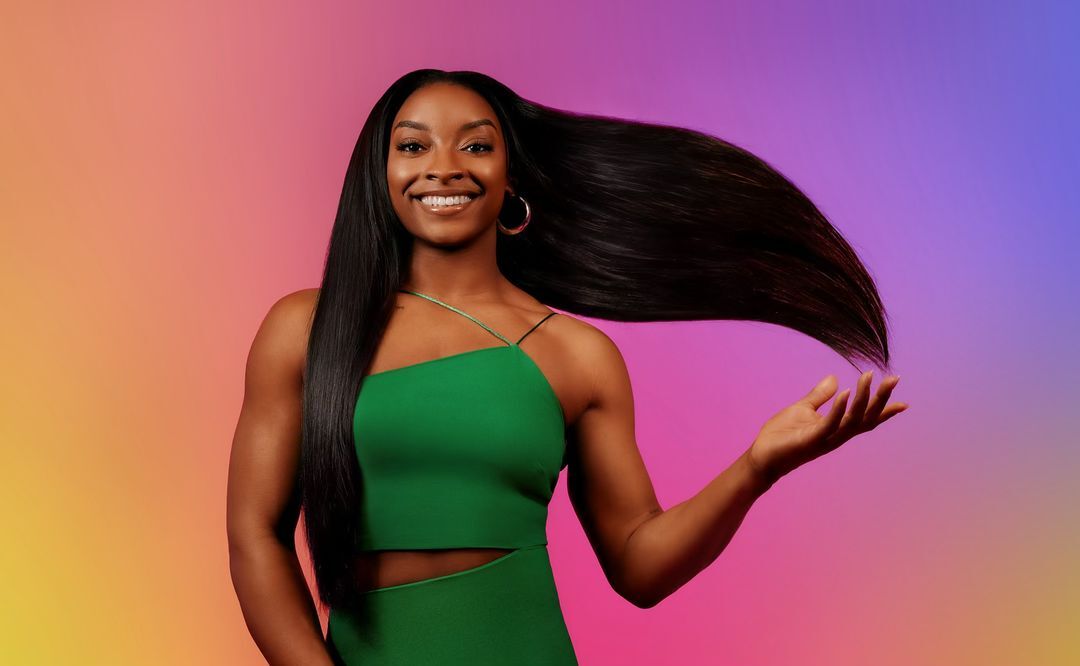 Smiling Black woman with long straight hair wearing a green dress in front of a vibrant colorful gradient background in a portrait shot.