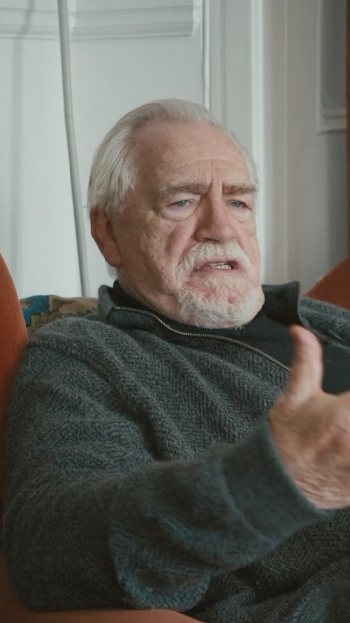 Close-up of an elderly man with gray hair in a gray sweater gesturing with his hand, indoors in front of a wall with a window.