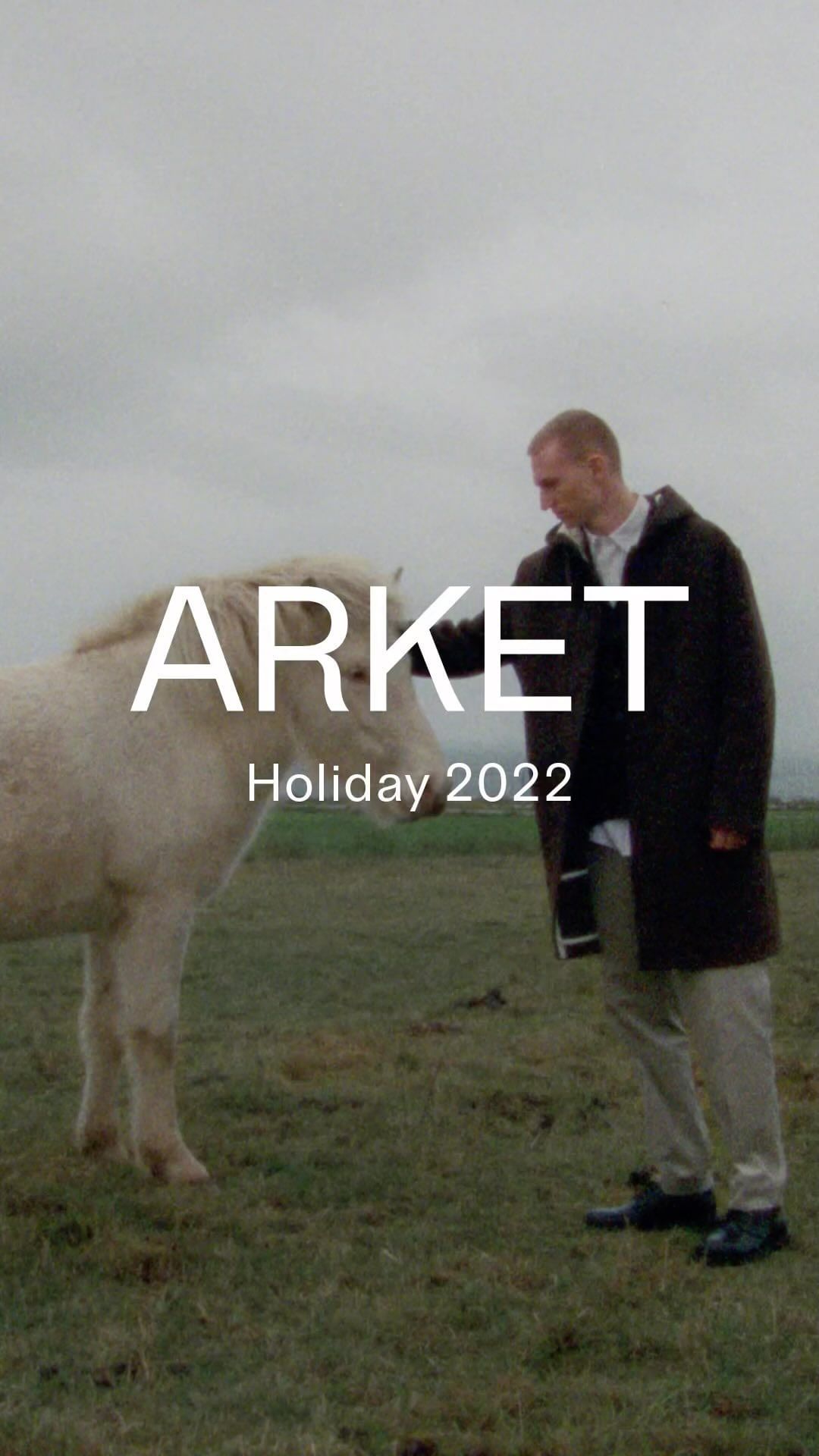 Man in coat stands next to a light-colored horse in a grassy field under overcast skies. Fashion editorial with animal.