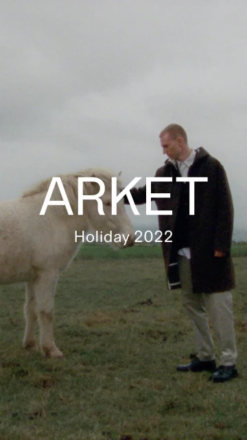 Man in coat stands next to a light-colored horse in a grassy field under overcast skies. Fashion editorial with animal.