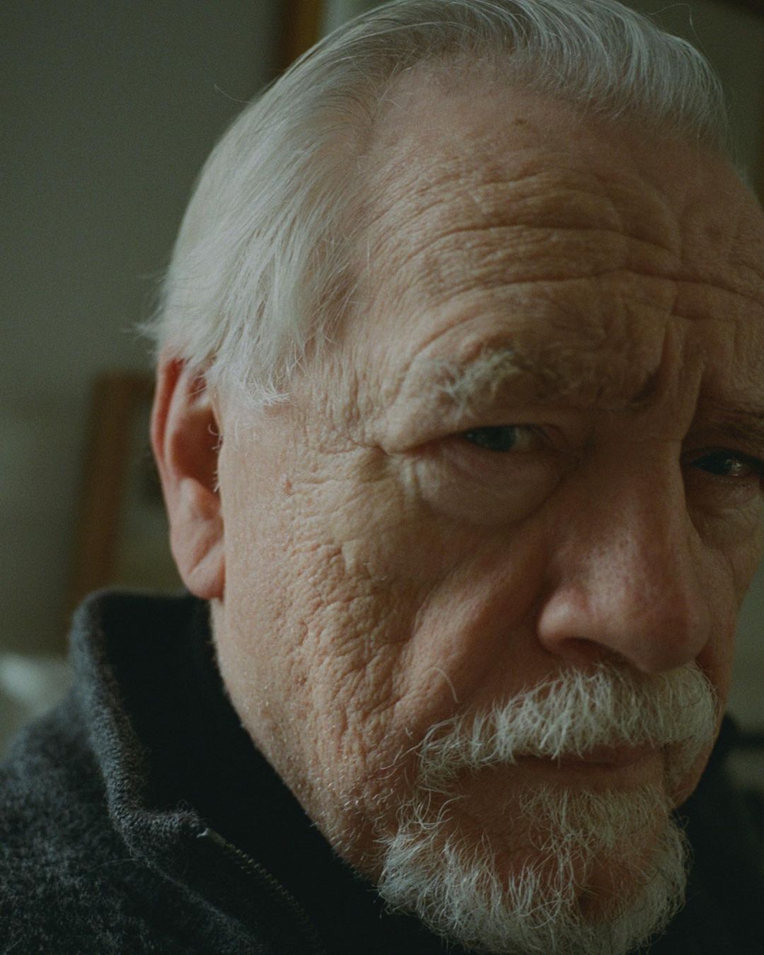 Close-up portrait of an elderly Caucasian man with white hair and a goatee, wearing a dark jacket against a neutral-toned background.