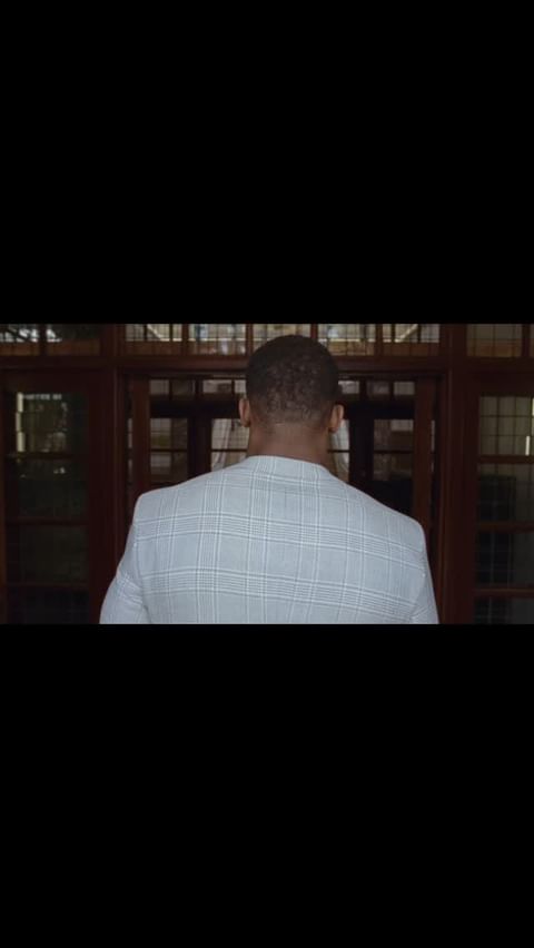 Back view of a Black adult male in a white checkered blazer entering a building with dark wood-framed glass doors.