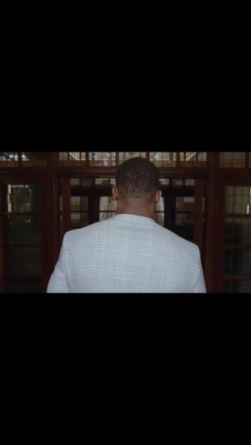 Back view of a Black adult male in a white checkered blazer entering a building with dark wood-framed glass doors.