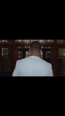 Back view of a Black adult male in a white checkered blazer entering a building with dark wood-framed glass doors.