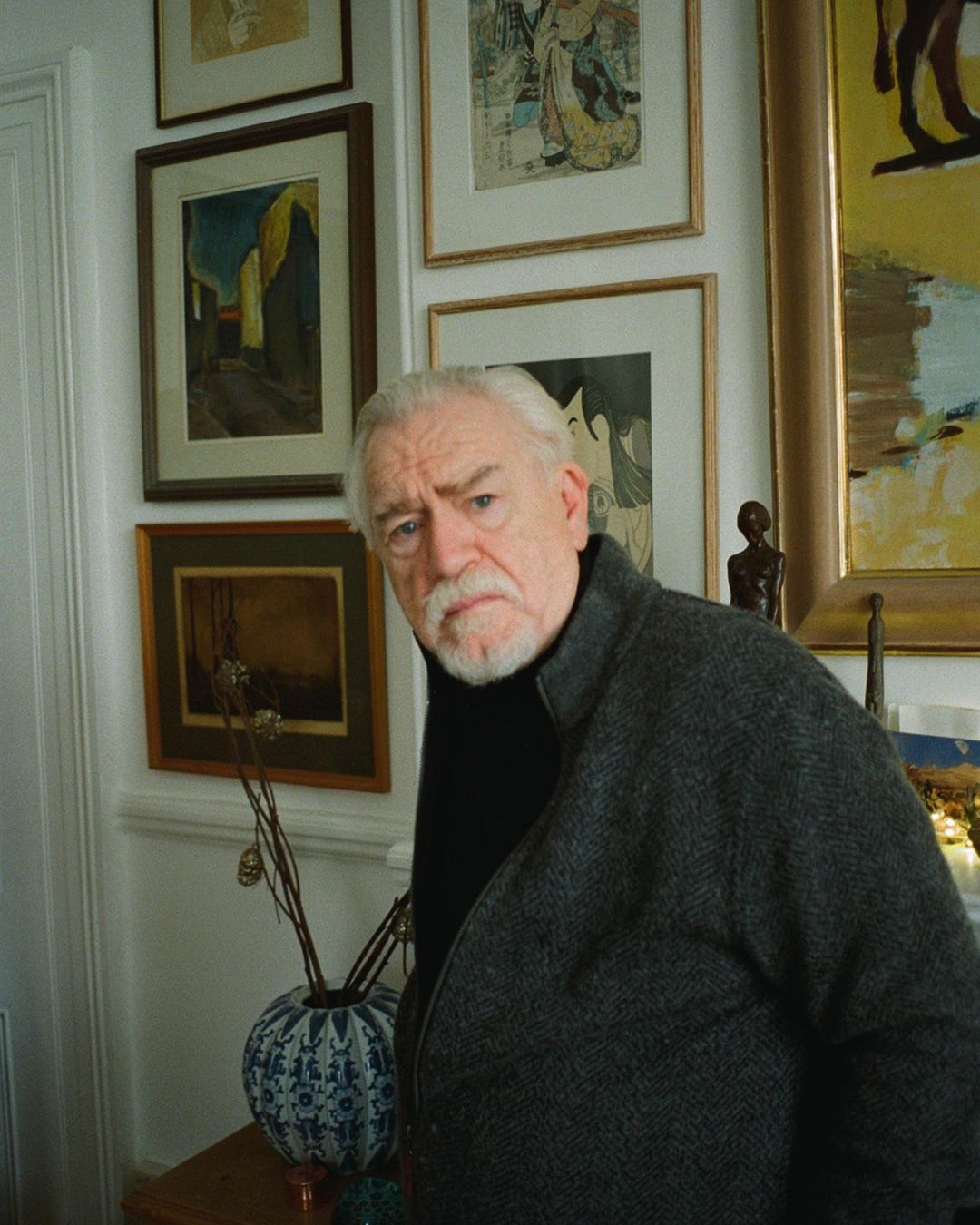 Portrait of an elderly man with a white beard wearing a grey jacket, standing indoors surrounded by framed art and a decorative vase.