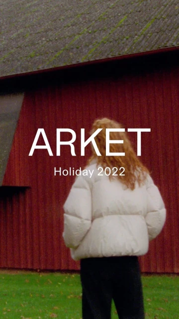 Woman in a white puffer jacket stands outdoors against a red barn with the brand ARKET and the text Holiday 2022 on the image.