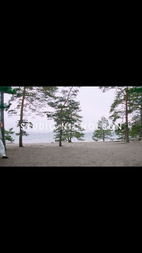Scenic outdoor shot showcasing a sandy path lined with tall pine trees and a serene lake under a cloudy sky, featuring the Studio Nicholson logo.