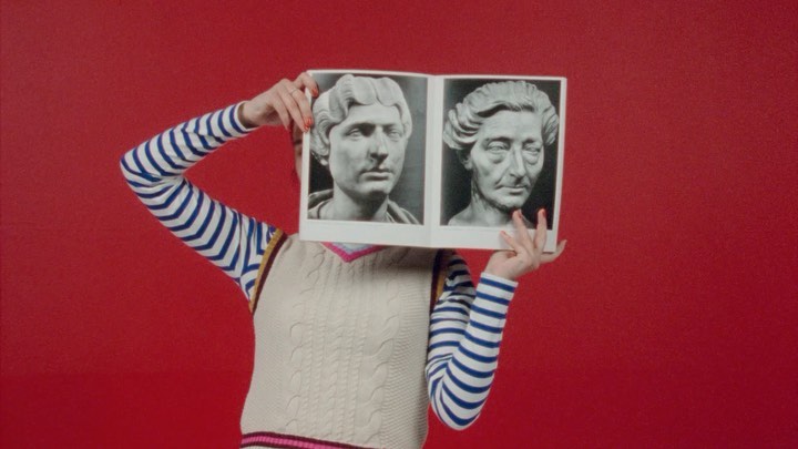 Person holding a book with classical sculpture faces against a red backdrop, wearing striped shirt and sweater vest in a studio setting.