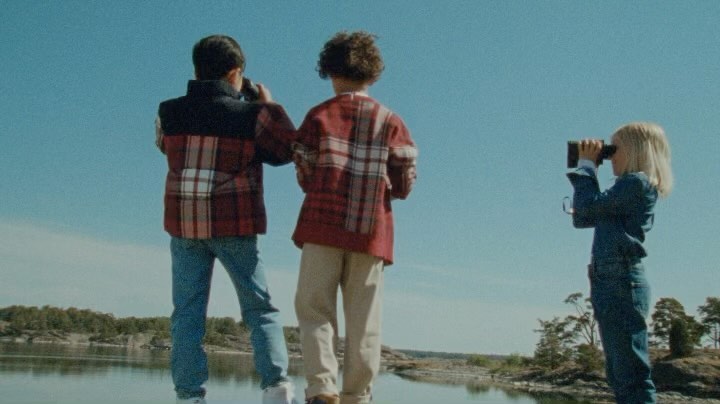Three children stand by a lake, looking through binoculars at the water and treeline against a clear blue sky in daylight.