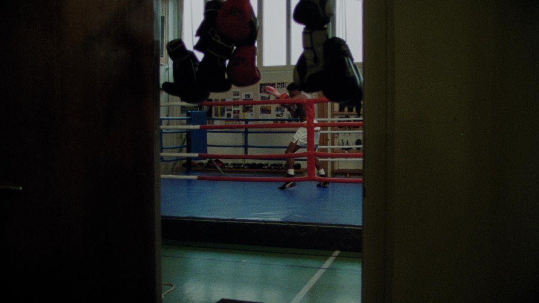 Boxer training in a boxing ring, framed by a partially open doorway. Red and black boxing gloves hanging overhead.