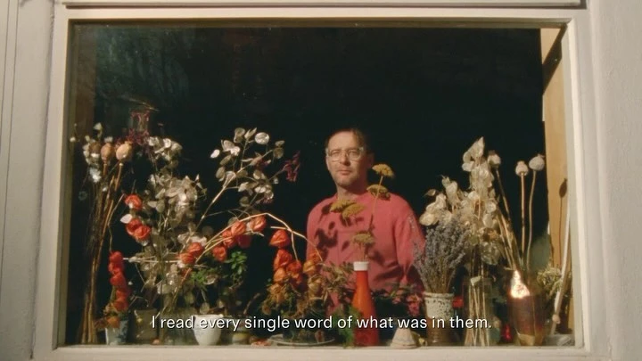 Man poses by a window display with red and white flowers in vases, bottle of ketchup, and a candle creating a floral-themed vignette.