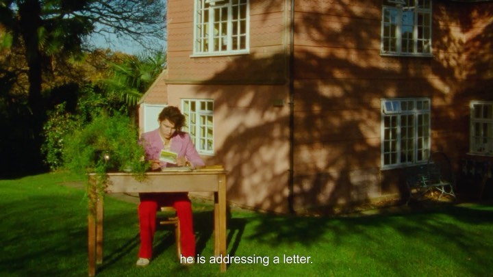 Male in a pink suit sits at a desk on a lawn, reading a card, with a red brick house and greenery visible in the background on a sunny day.