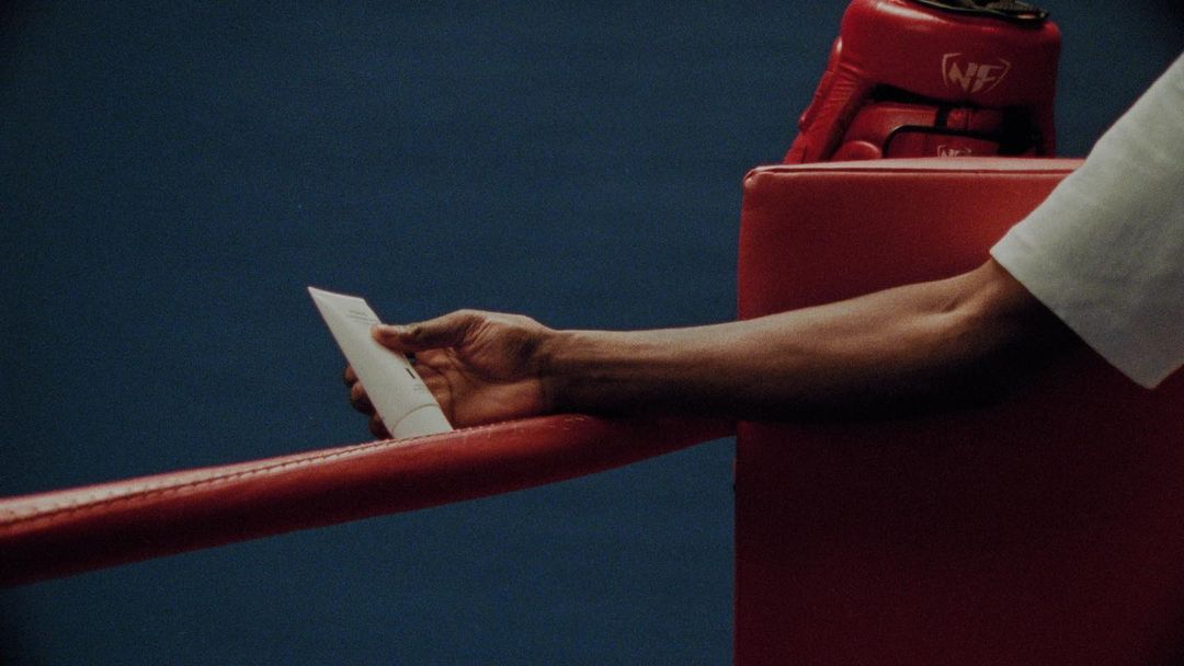 Close-up of an arm holding a white tube, set against a red boxing ring rope and a dark blue wall, with a red boxing helmet.