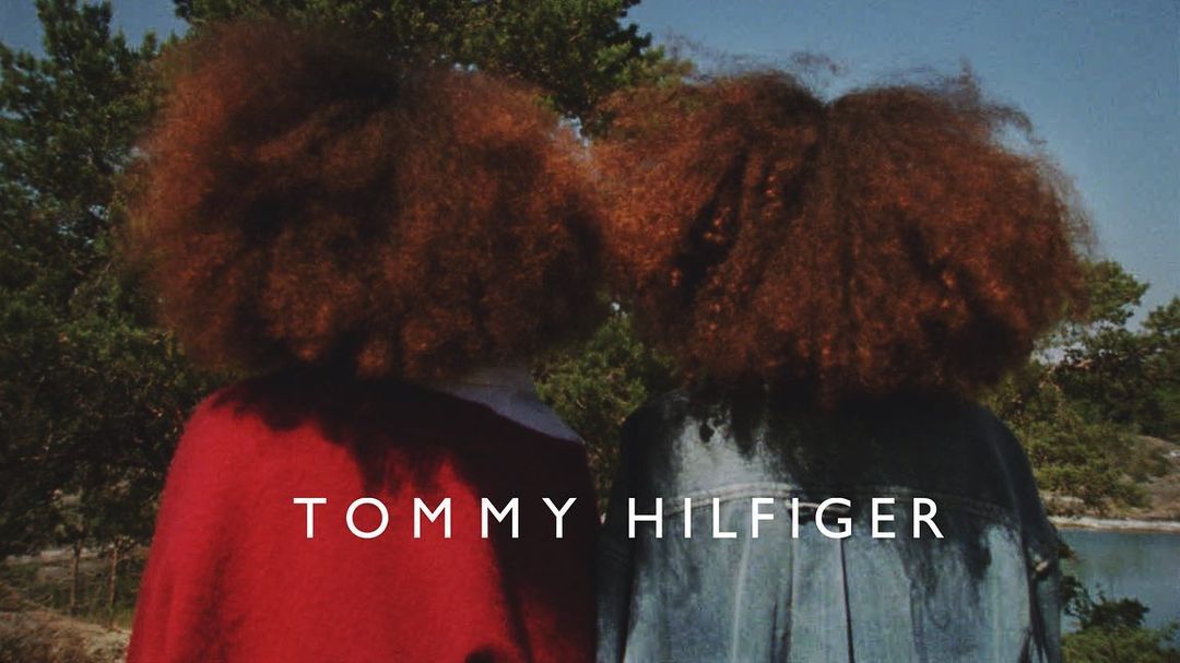 Two people with auburn afro hairstyles and wearing jackets, standing outside near greenery under blue sky, Tommy Hilfiger text overlay.
