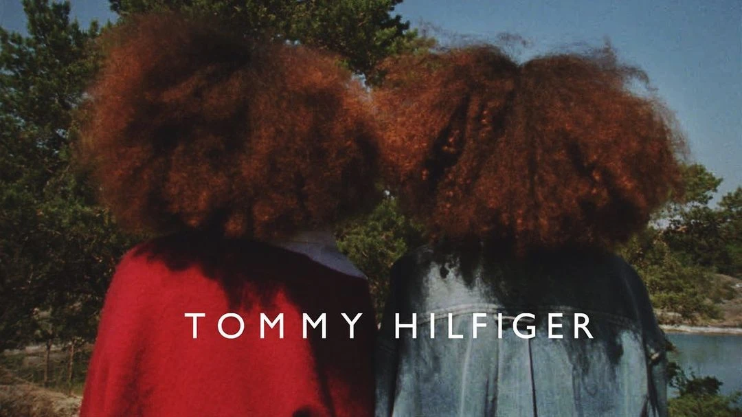 Two people with auburn afro hairstyles and wearing jackets, standing outside near greenery under blue sky, Tommy Hilfiger text overlay.