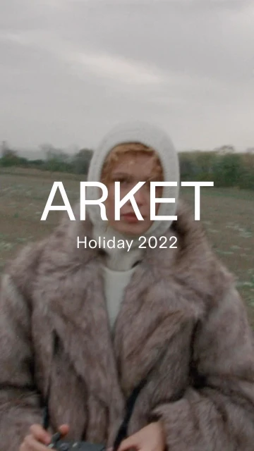 A person wearing a fur coat and hoodie stands in a field on a cloudy day. Text overlays the image including brand and holiday information.