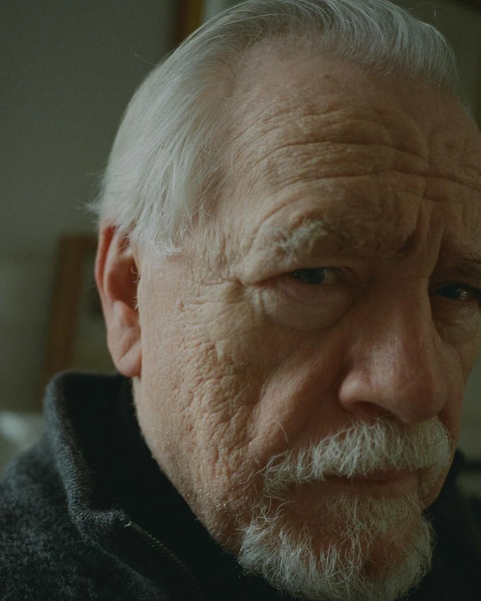 Close-up portrait of an elderly white man with a white goatee and wrinkled skin wearing a dark grey textured sweater.