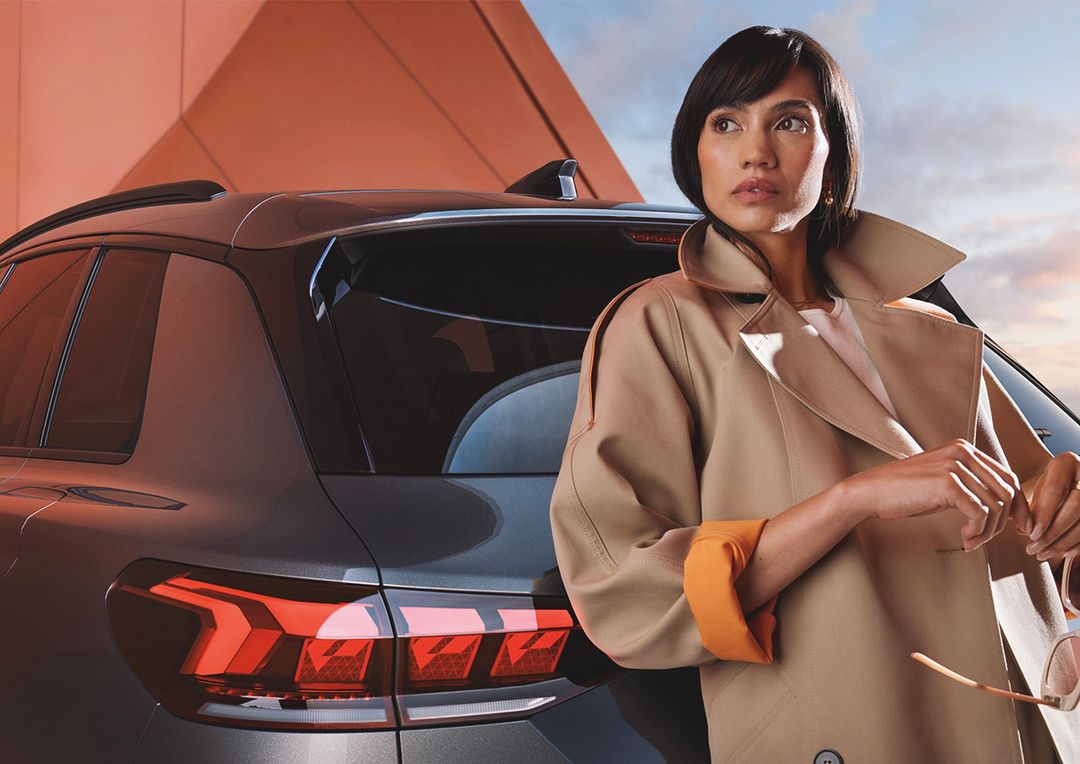 Adult woman in trench coat poses next to a grey SUV car with striking red tail lights; modern architecture and sky at dusk visible.