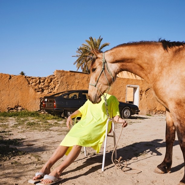 Conceptual outdoor shot with a horse standing near a person in a yellow shirt sitting on a chair, vintage car, and adobe building.