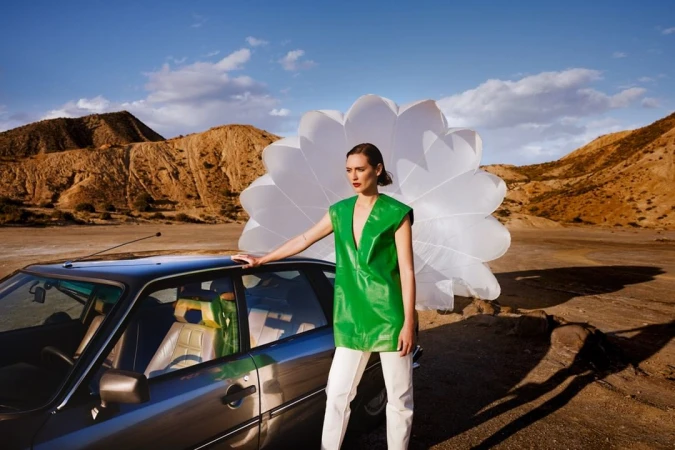 Model in green top and white pants leaning on a car with a large white pleated prop in a desert landscape