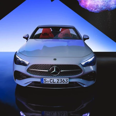 Front-angle studio shot of a white Mercedes-Benz, showcasing its grille, headlights, and sleek design, with a blue and black abstract backdrop.