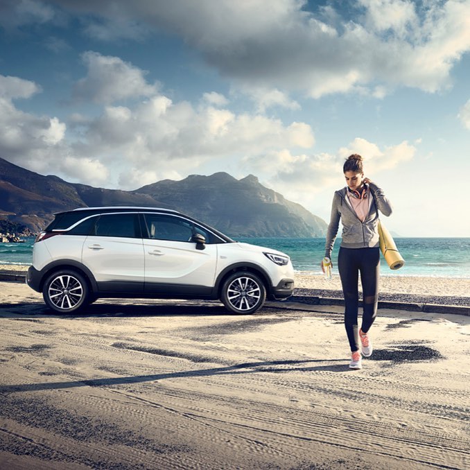 White crossover SUV parked near a woman with a yoga mat at a beach, with mountains and a cloudy sky in the background on a sunny day.