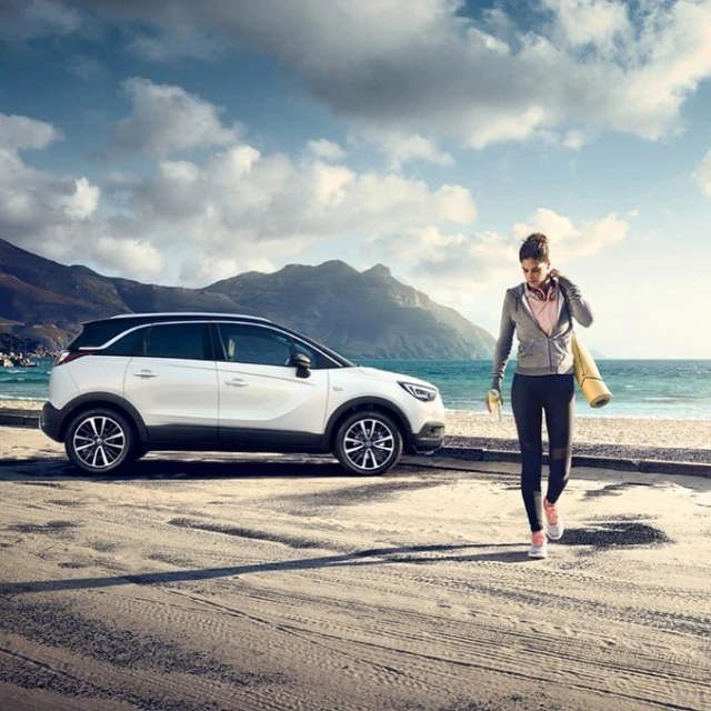 White crossover SUV parked near a woman with a yoga mat at a beach, with mountains and a cloudy sky in the background on a sunny day.
