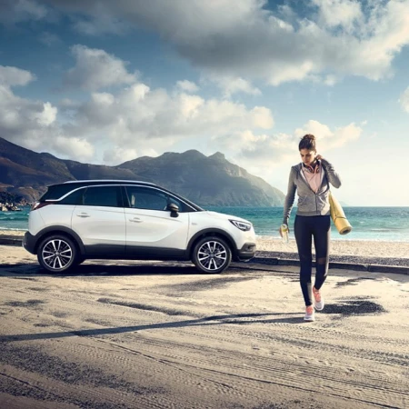 White crossover SUV parked near a woman with a yoga mat at a beach, with mountains and a cloudy sky in the background on a sunny day.