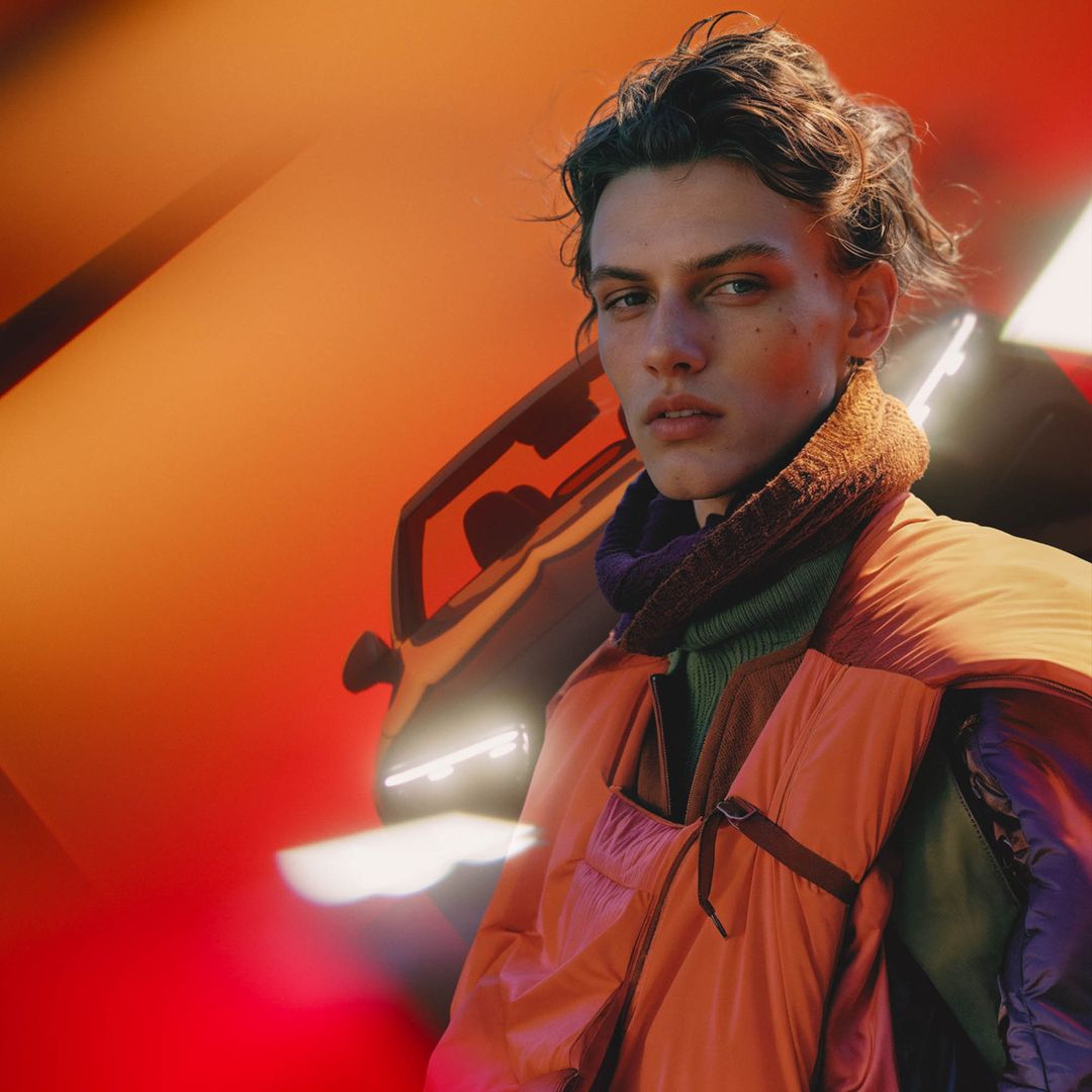 Portrait of a young adult male with an orange puffer vest standing near the front of a car, with soft lighting and a blurred background.