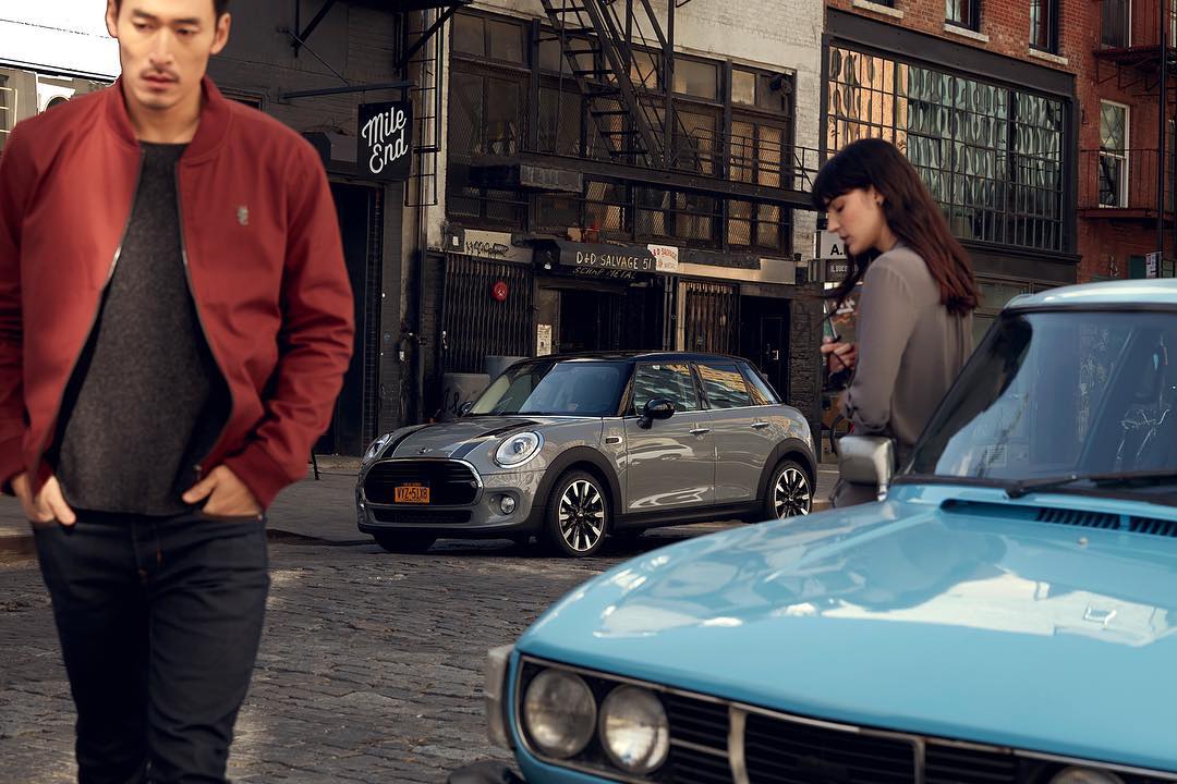 Automotive advertisement featuring a grey Mini Cooper parked on a city street, an adult male walking, and an adult female by a blue car.