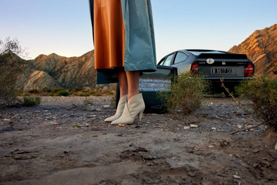 Outdoor fashion shot featuring adult female legs with light-colored heeled boots, orange skirt, and a teal coat with a vintage car and mountains.