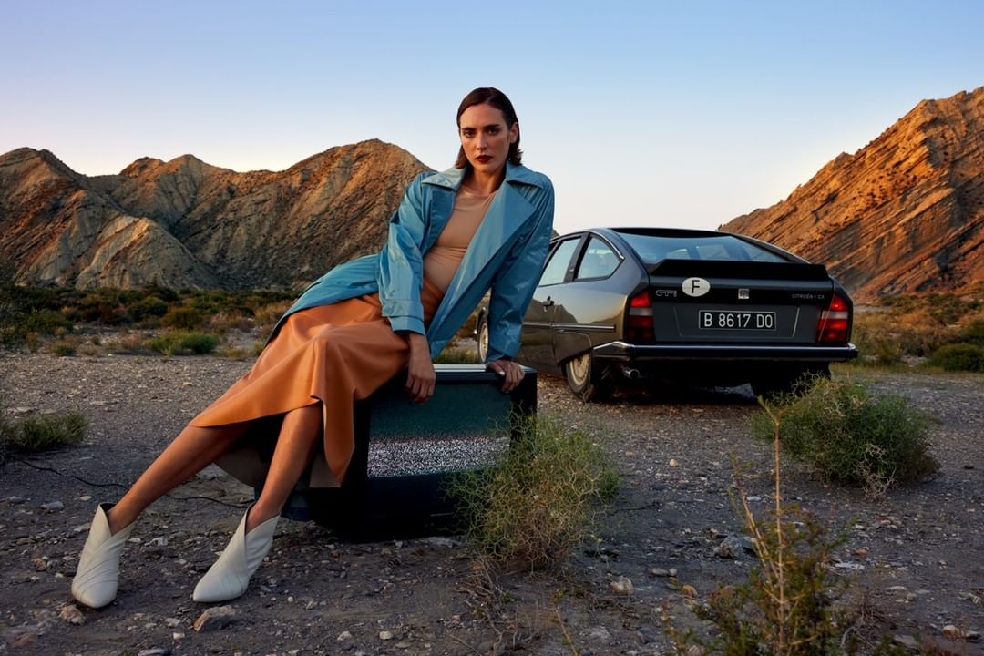 A woman in a trench coat sits on an old television set, posed next to a vintage car with a desert mountain range background at dusk.