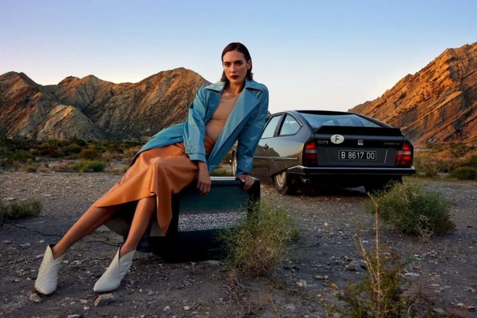 A woman in a trench coat sits on an old television set, posed next to a vintage car with a desert mountain range background at dusk.