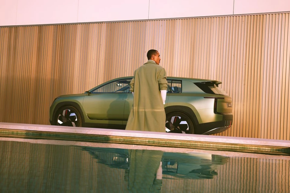 Stylish electric SUV car in olive green, parked by an outdoor water feature with a person in a long coat standing near the vehicle.