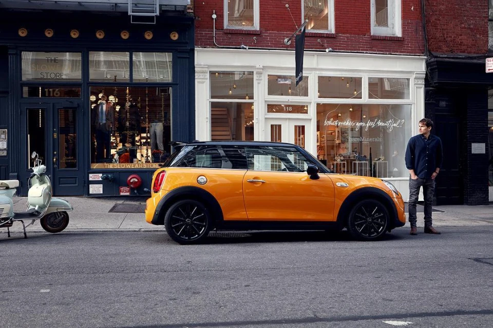 A yellow Mini Cooper is parked on a city street next to a man, with buildings and storefronts in the background on a sunny day.