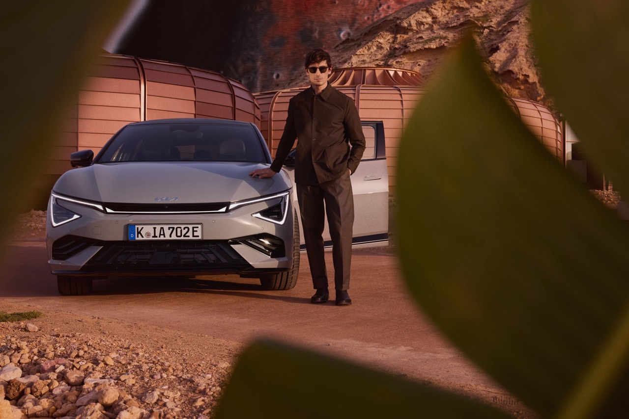 A light grey Kia EV6 electric car with an adult male model posing in front of an organic architectural structure in an outdoor setting.