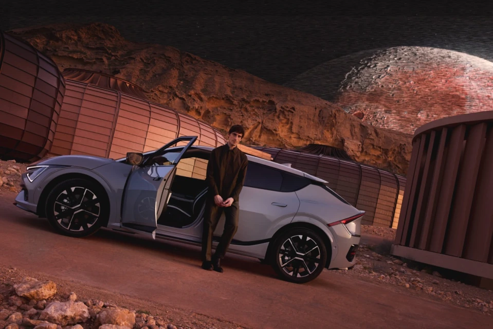 Man stands beside a light-grey electric car on a reddish desert road, set against a backdrop of unusual architecture and geological formations.