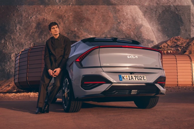 A gray Kia EV6 car stands with an adult male leaning against it, set against a planetary background with space structures.