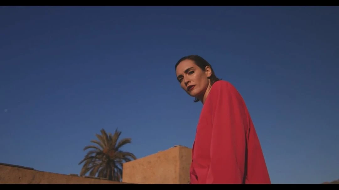 Adult female poses outdoors in a red coat against a blue sky and palm tree. Fashion editorial shot. Minimalist aesthetic, saturated color.