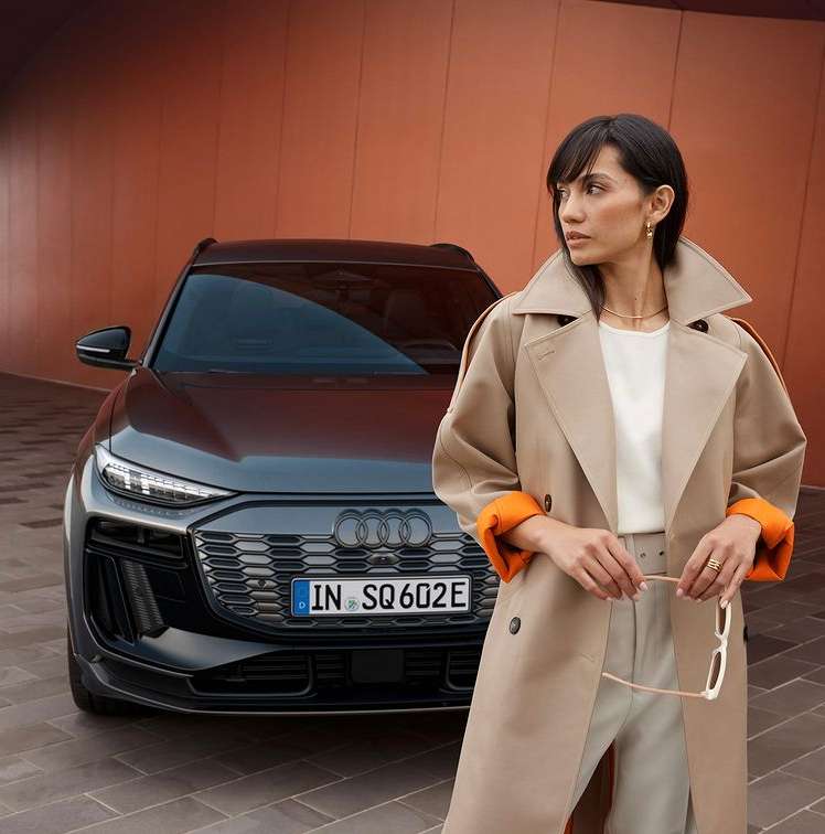 A woman in a trench coat stands in front of a dark grey Audi SUV with an orange wall in the background, showcasing automotive fashion.