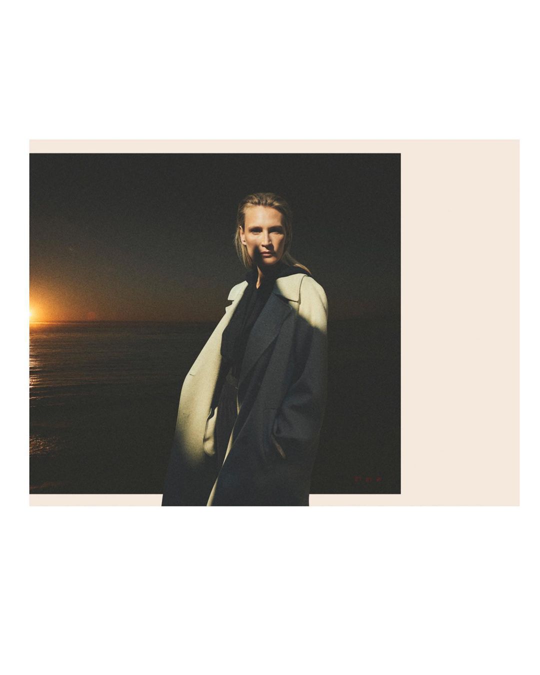 A woman in a trench coat stands against a dark coastal backdrop as the sun sets over the ocean. Editorial fashion shot.