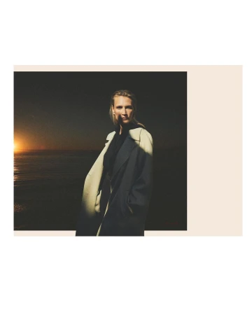 A woman in a trench coat stands against a dark coastal backdrop as the sun sets over the ocean. Editorial fashion shot.