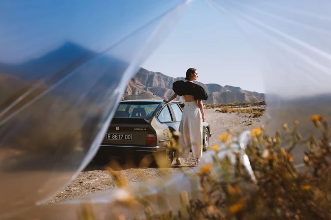 A woman leaning on a vintage car parked on a dusty desert road framed by sheer fabric and yellow wildflowers.