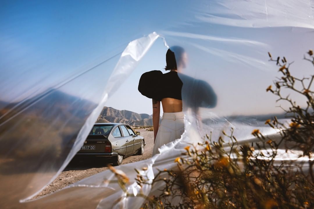 Fashion shot of a woman in a black top and white pants, partially obscured by plastic, with a car in a desert landscape.