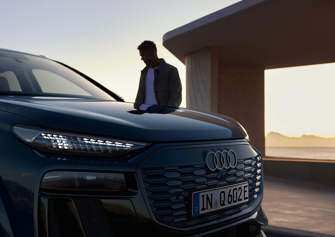 A dark blue Audi Q6 e-tron parked outside with an adult male standing near the hood of the car during daytime with a coastal backdrop.