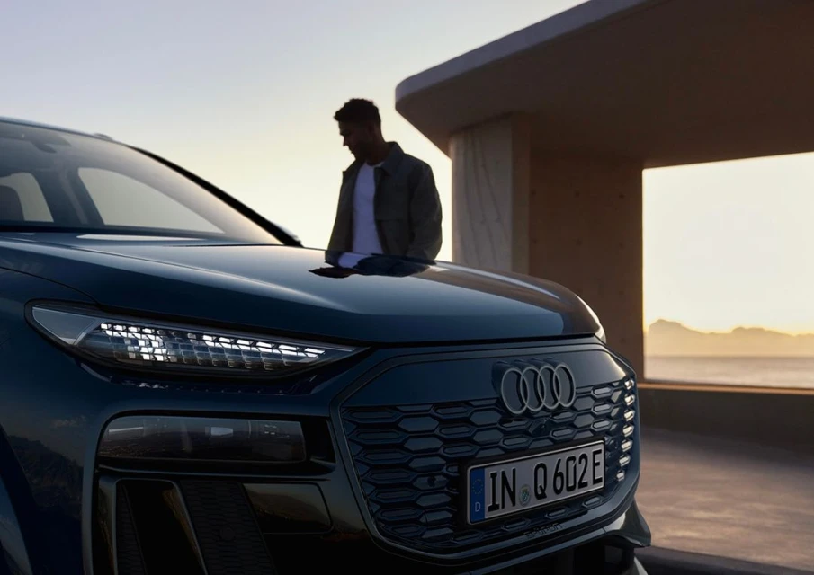 A dark blue Audi Q6 e-tron parked outside with an adult male standing near the hood of the car during daytime with a coastal backdrop.
